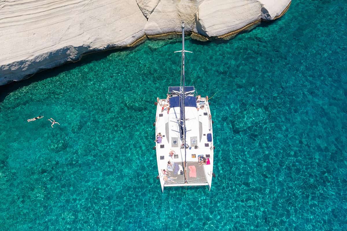 Activities in Milos: a sailing catamaran anchored in turquoise water below the white rocks of Kleftiko, with swimmers nearby