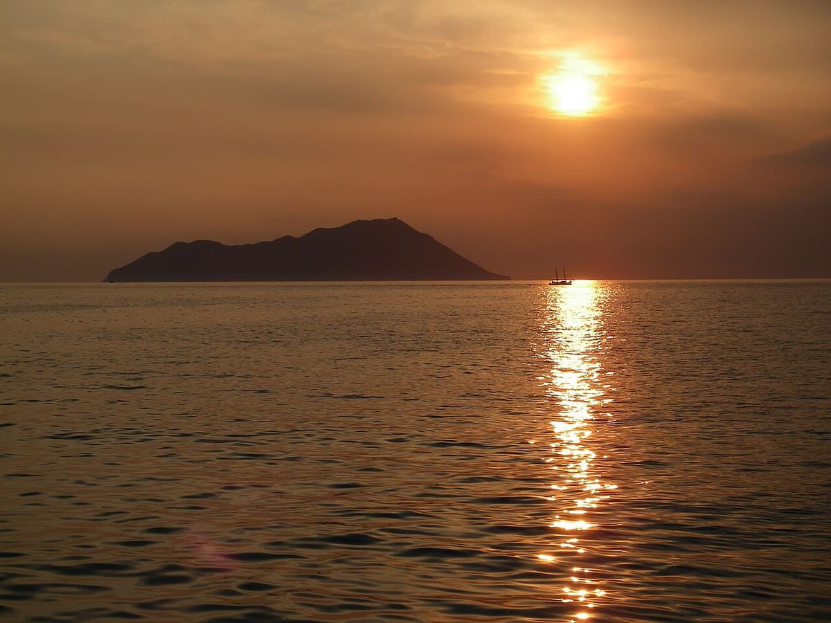 Antimilos: the dark outline of the islet against an orange sunset sky, with the sun reflected on the calm sea