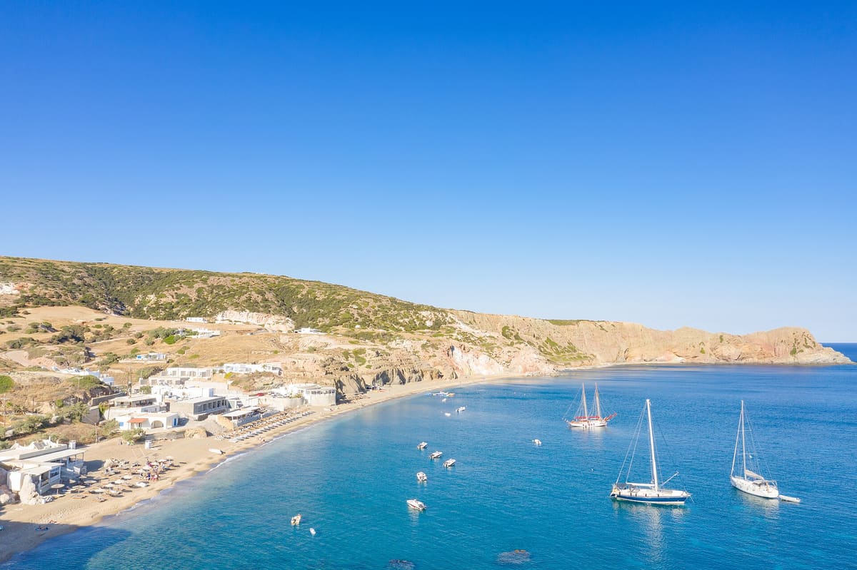 Coffee and drinks in Paleochori: the beach of Paleochori from the air, with sailing boats in the clear water, umbrellas on the sand and the coloured cliffs behind
