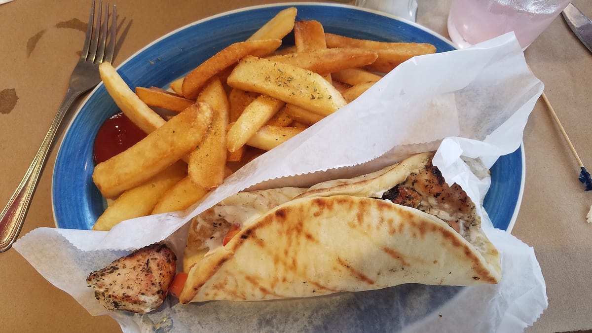 Food in Triovasalos: a grilled souvlaki pita wrapped in paper, served with fried potatoes on a blue plate