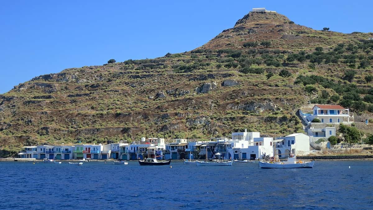 Kayak to Klima Milos: the painted boathouses of Klima along the shore below a terraced hill, seen from a small boat on the gulf