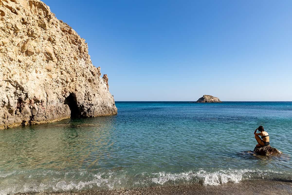Kayak Tsigrado: clear shallow water at Tsigrado below a tall pale rock with a small cave, an islet on the horizon