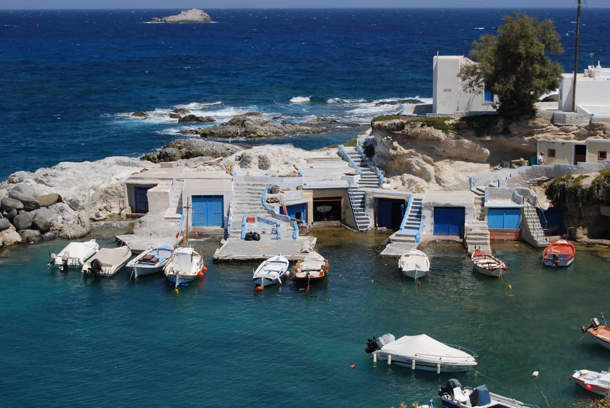 Mandrakia: whitewashed boathouses with blue doors carved into the rock, small fishing boats moored in the green water of the cove