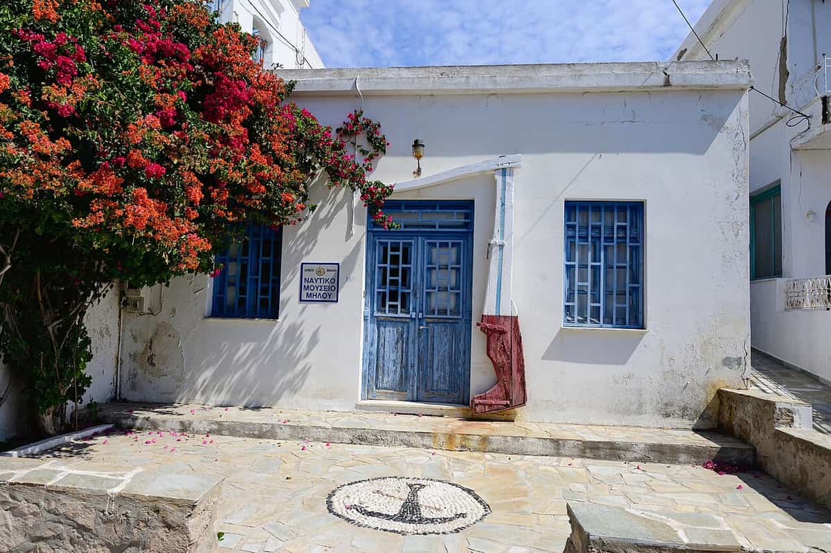 Naval museum of Milos: the whitewashed museum house in Adamantas with blue doors and windows, a ship's rudder by the entrance and an anchor in the pebble mosaic