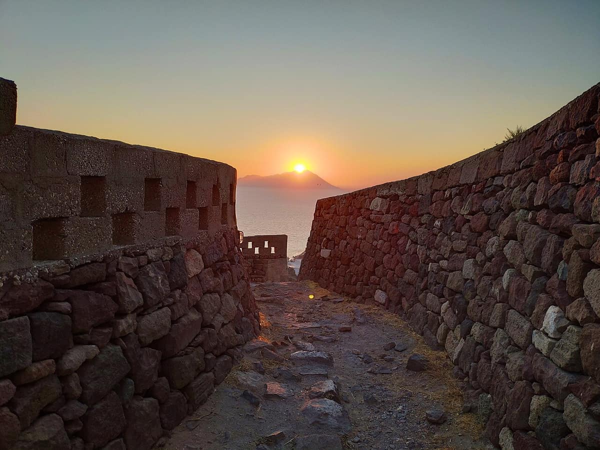 Milos nightlife: the sun setting over the sea between the old stone walls on the castle hill above Plaka