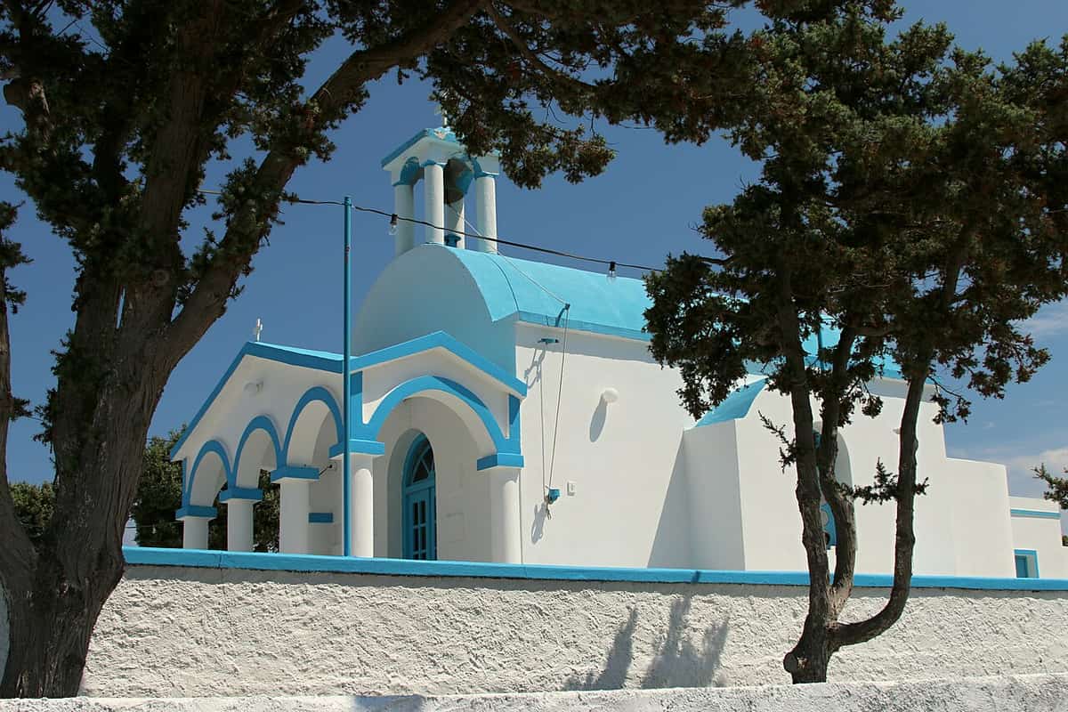 Pollonia: the white church of Agios Nikolaos with blue domes and arches behind tamarisk trees in the fishing village on Milos