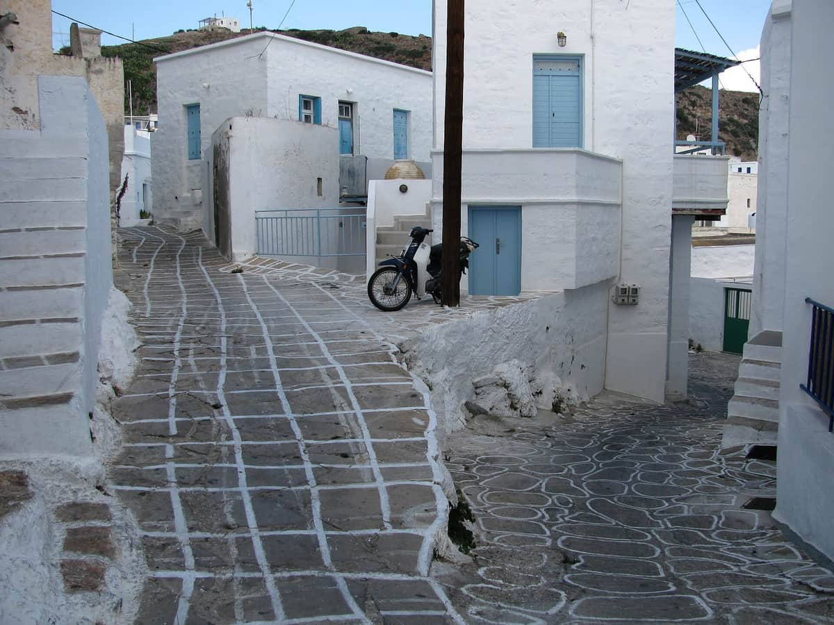 Rent a car Kimolos: a paved lane with white-lined stones between whitewashed houses in Chorio, with a scooter parked by a door