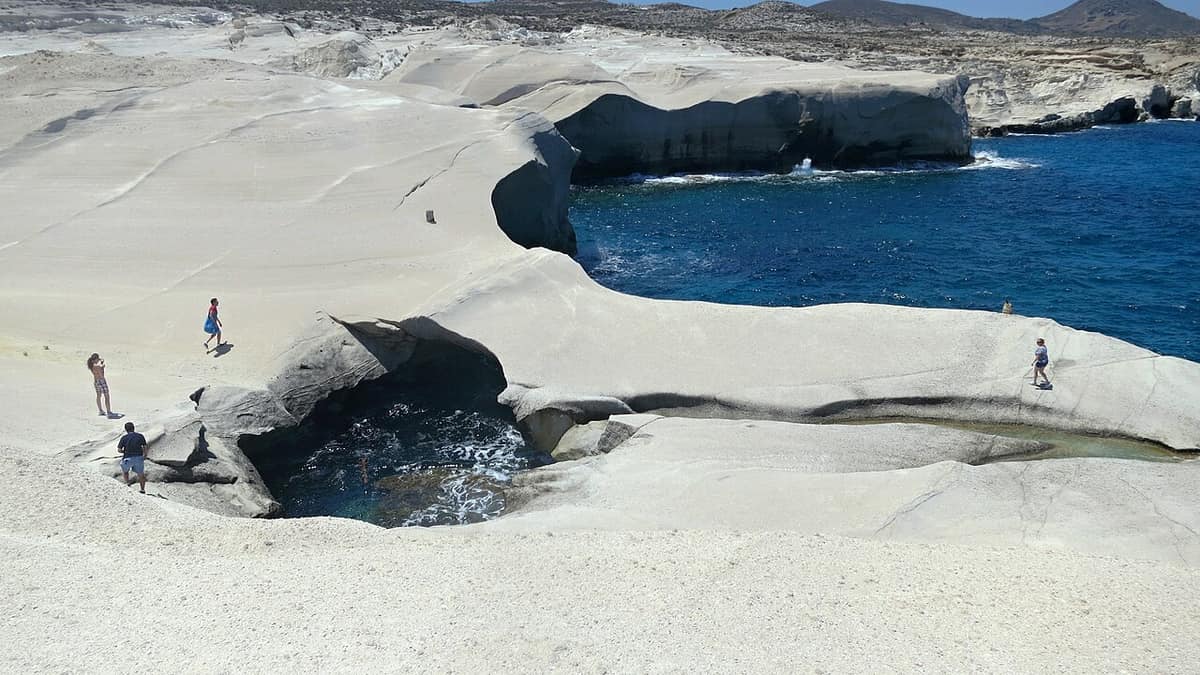 Sarakiniko beach: people standing on white rock ledges above a small sea arch and deep blue water