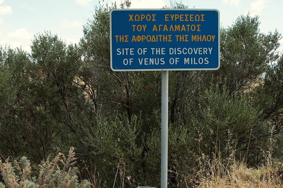 Venus de Milo find spot: a blue sign in Greek and English marking the site of the discovery on Milos, among olive trees