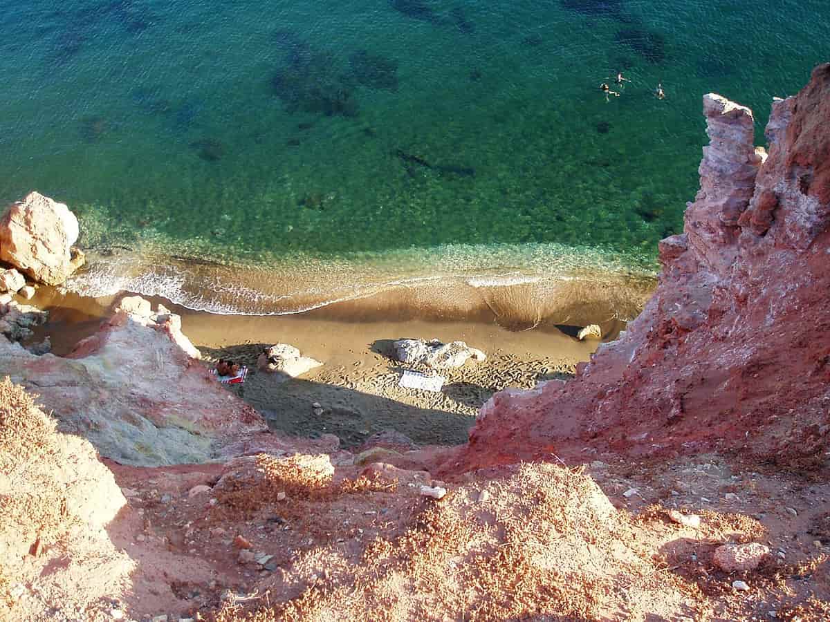 Milos with kids: clear shallow water over a sandy beach below red and white rocks at Paleochori
