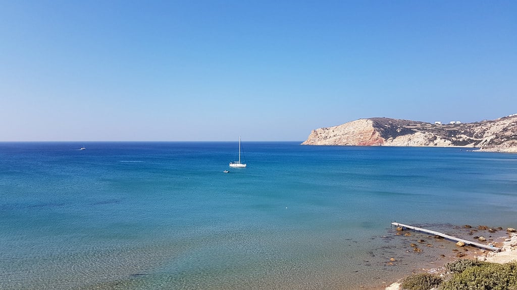 Ιστιοπλοΐα Μήλος: ένα ιστιοπλοϊκό αγκυροβολημένο σε γαλάζιο κόλπο, με λευκά βράχια στο βάθος