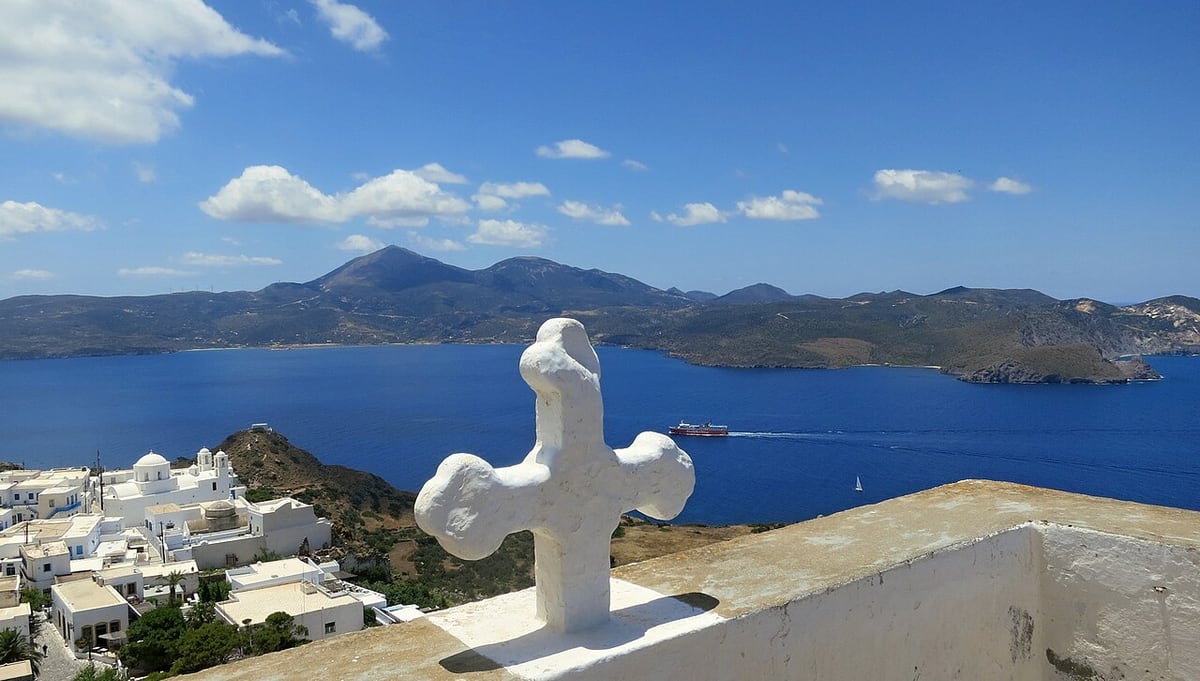 Κάστρο Πλάκας: λευκός σταυρός πάνω σε τοίχο στην κορυφή, με τα σπίτια της Πλάκας, τον κόλπο και τα βουνά της Μήλου στο βάθος