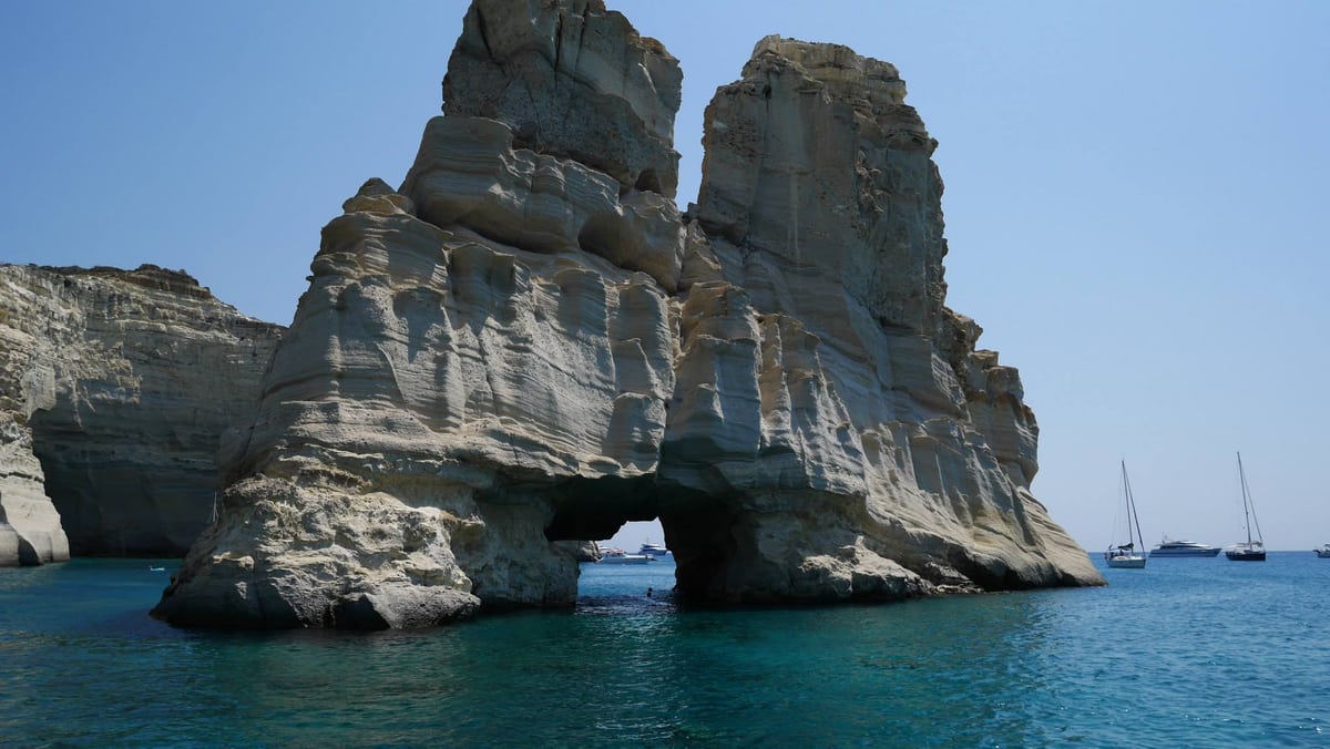 Λευκοί ηφαιστειακοί βράχοι του Κλέφτικου μέσα στη θάλασσα, με ιστιοπλοϊκά αγκυροβολημένα πίσω τους