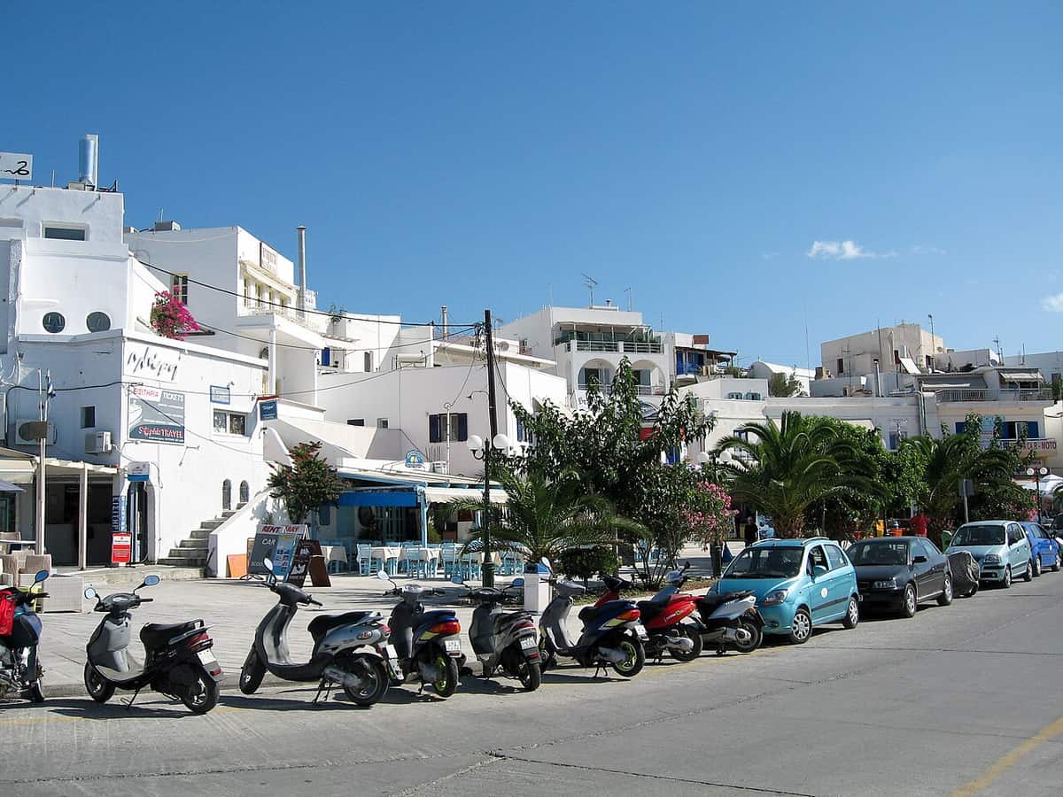Λεωφορεία Μήλος: ο παραλιακός δρόμος του Αδάμαντα με λευκά σπίτια, φοίνικες, αυτοκίνητα και μηχανάκια