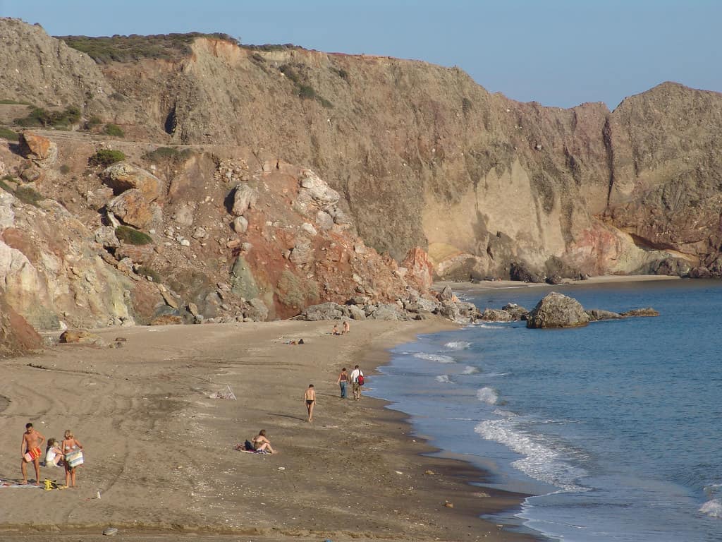 Nude beach Milos: a long sandy shore under tall red and brown cliffs at Paleochori