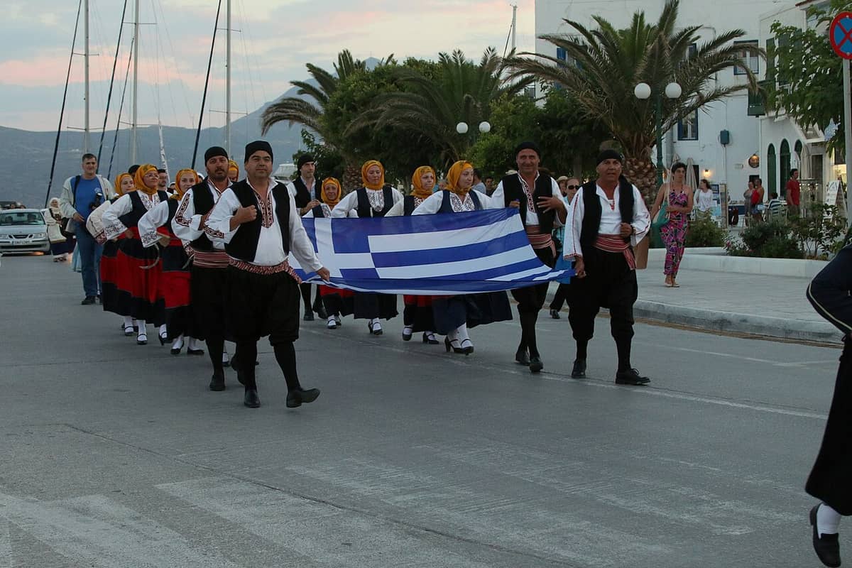 Νυχτερινή ζωή Μήλος: χορευτές με παραδοσιακές στολές και ελληνική σημαία περνούν από την παραλία του Αδάμαντα το σούρουπο