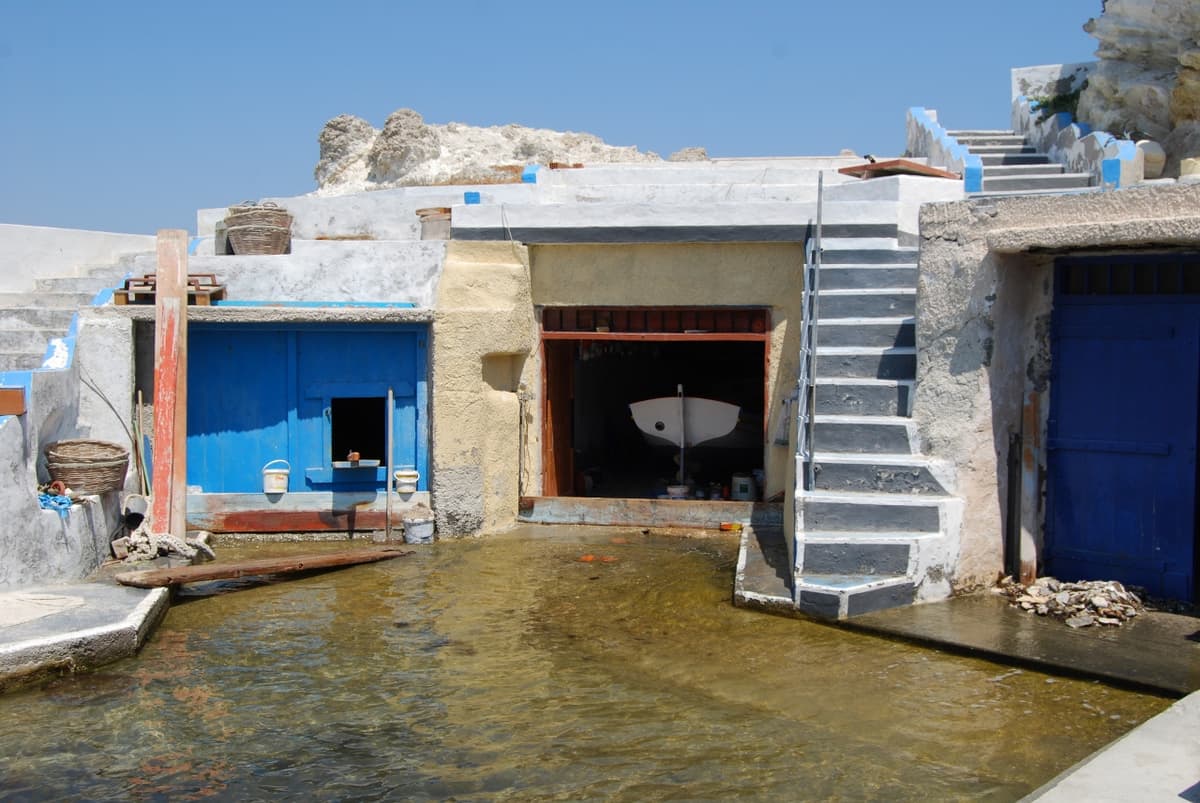 Salty Breeze Boathouse Μήλος: σύρμα σκαμμένο στον βράχο με μπλε πόρτες, βάρκα στο ισόγειο και σκάλα προς τον όροφο