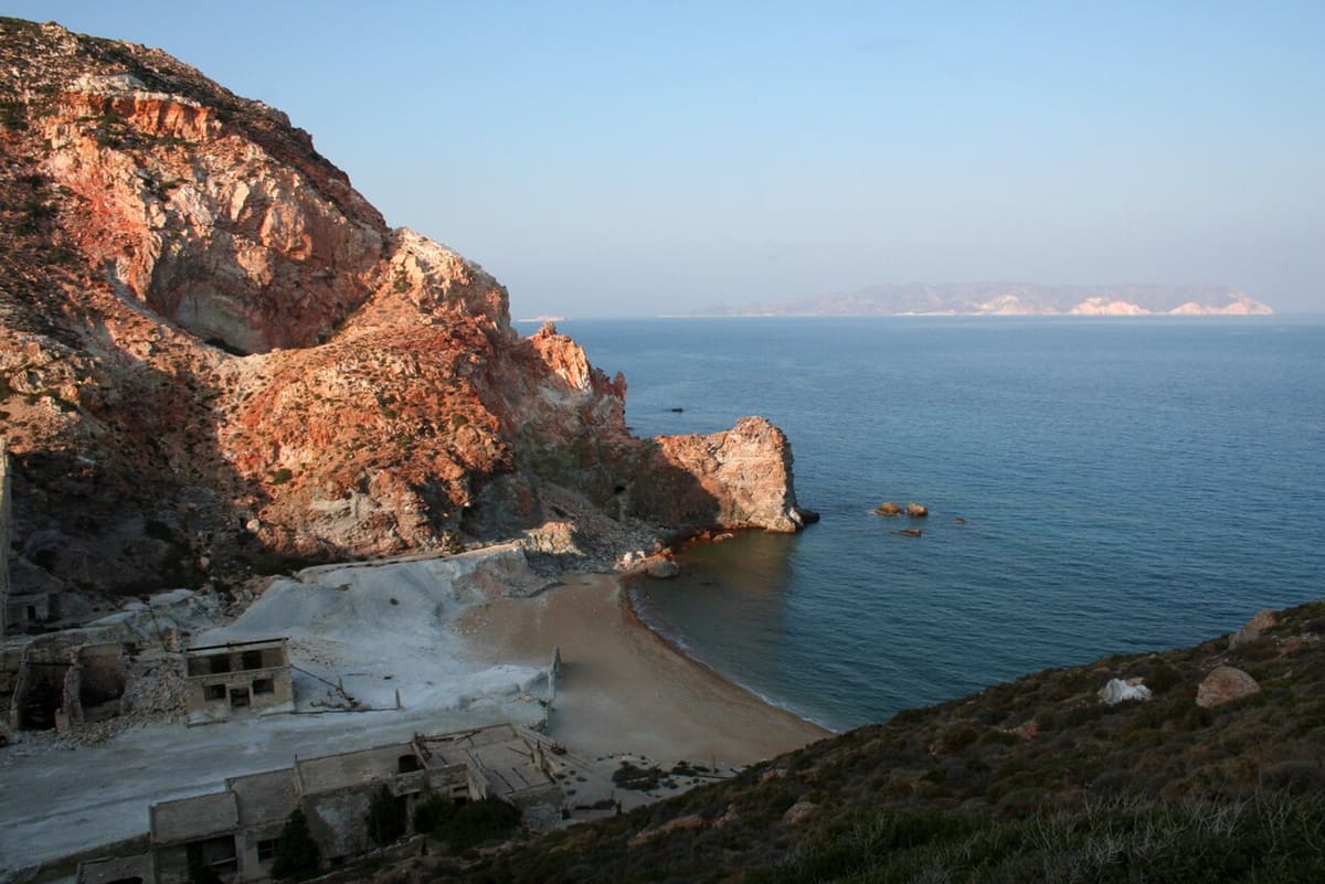 Θειωρυχεία: κόκκινοι και πορτοκαλί βράχοι πάνω από μικρή αμμουδιά, με ερείπια του ορυχείου και ήρεμη θάλασσα