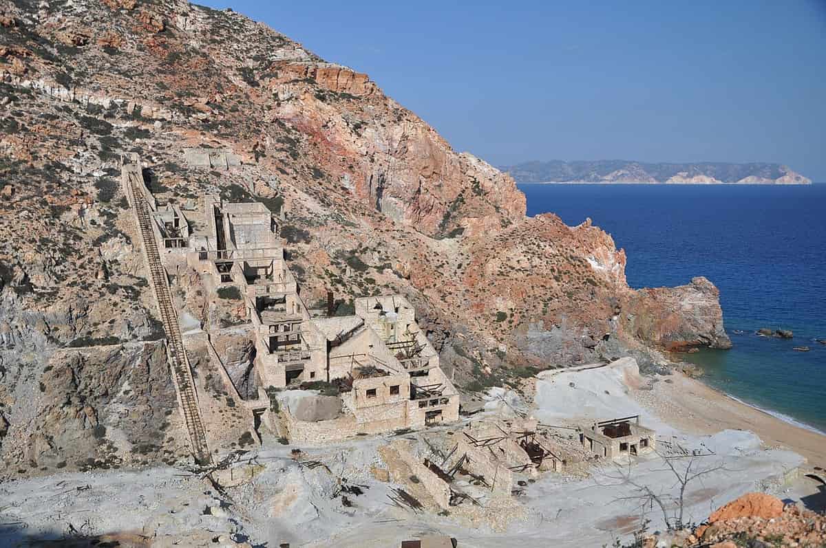 Θειωρυχεία Μήλου: τα εγκαταλειμμένα κτίρια του ορυχείου θείου πάνω από την αμμουδιά, με κόκκινους και λευκούς βράχους και ανοιχτή θάλασσα